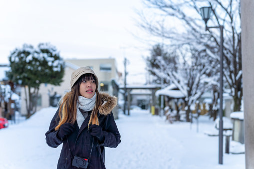 Young Asian woman in winter coat walking on city street covered in snow in snowy day evening. Attractive girl enjoy urban outdoor lifestyle travel in Japan on holiday vacation in winter season.