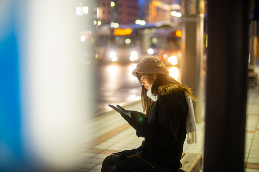 Asian woman working on digital tablet and listening to the music on earphones during waiting for bus at night. Attractive girl enjoy urban outdoor lifestyle travel in the city with wireless technology