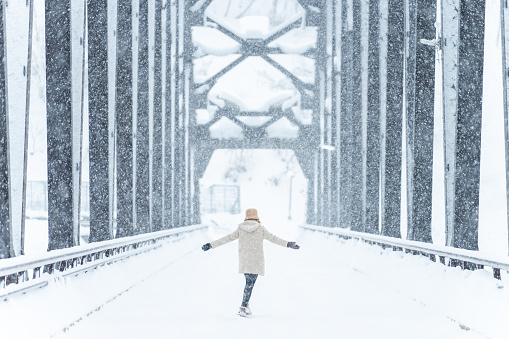 Young Asian woman walking on steel bridge over the river between the mountain and small town covered in snow. Attractive girl enjoy and fun travel local village on winter holiday vacation in snowy day