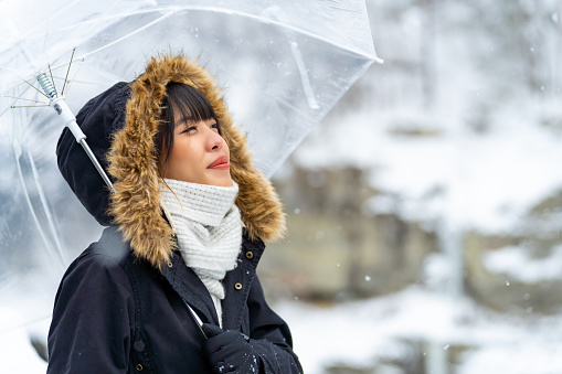 Young Asian woman in winter coat holding umbrella walking down footpath on forest mountain near small town in snow day. Attractive girl tourist solo travel local village in Japan on winter holiday vacation.