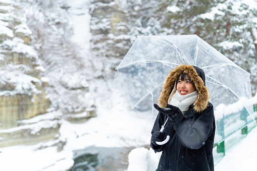 Young Asian woman in winter coat holding umbrella walking down footpath on forest mountain near small town in snow day. Attractive girl tourist solo travel local village in Japan on winter holiday vacation.