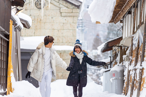 Young Asian couple holding hands walking together during travel Ouchi-juku village in Fukushima prefecture covered in snow. Man and woman enjoy outdoor lifestyle travel Japan on winter holiday vacation.
