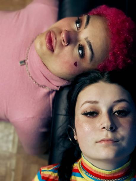 Two diverse, gender neutral, mixed race teens lying on a sofa together indoors. The girls are wearing pink and rainbow colored clothing. The girls represent mixed races, gender neutrality, and they would like to grow up without labels. The girls are bonding and relaxing indoors.