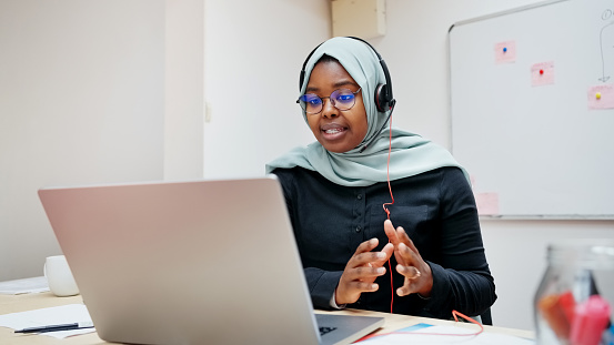Muslim woman teacher in hijab with headset tutoring students online using a laptop and webcam from her office