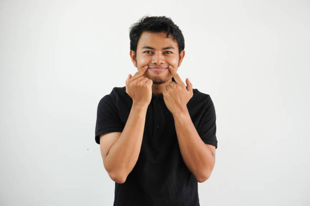 istock young asian man in black polo t shirt, white studio backdrop doubting between two options. force a smile with a finger. 1586665077