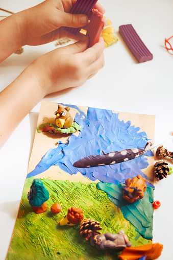Child smearing colorful plasticine on cardboard and creating fairy tale card with cartoon animals, using nature details also, dry flowers, cone, leaves