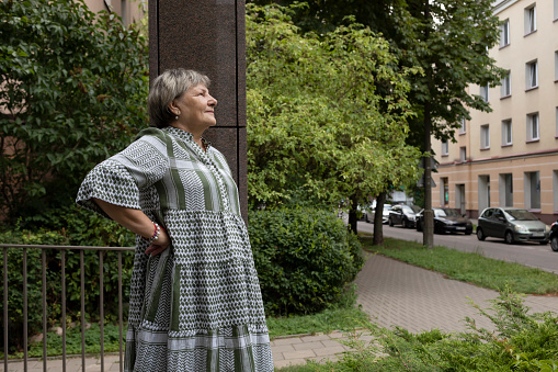 Real Smiling Female Woman In Yard Of Modern Building. Green Trees Around. Copy Space For Text. Pleased Elderly 70s Carefree, Full of Hope Beautiful Granny Looking Away Horizontal Plane