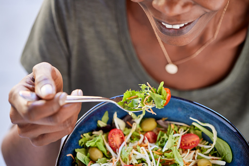 Close up of happy black woman eating healthy salad with green fresh ingredients. Smiling vegetarian woman holds bowl of fresh salad while eating tomatoes and carrot with fork. Detail of beautiful african american lady eating vegan food at home during lunch.