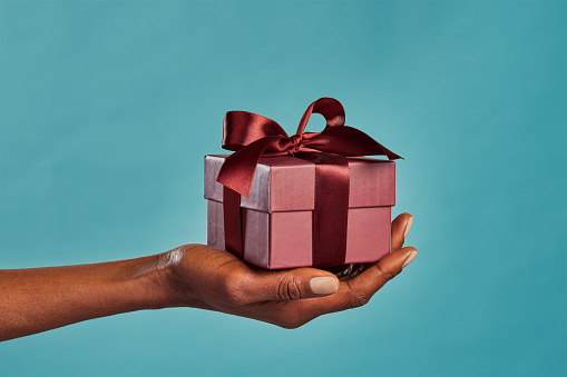 Close up shot of female hand holding a small gift wrapped with elegant brown ribbon isolated against blue background. Small craft gift in the hands of african american woman. Closeup of black woman holding elegant little box in hand for Christmas.