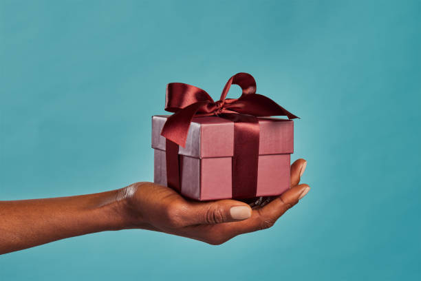 Black female hand holding gift box on blue Close up shot of female hand holding a small gift wrapped with elegant brown ribbon isolated against blue background. Small craft gift in the hands of african american woman. Closeup of black woman holding elegant little box in hand for Christmas. hand holding gift stock pictures, royalty-free photos & images