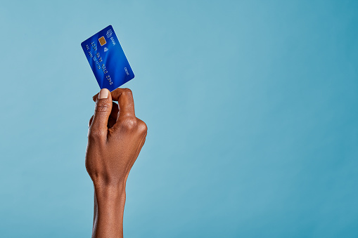 Close up of african woman hand holding bank credit card to make a payment with copy space. Black woman hand holding credit card for financial transaction or contact less payment isolated on blue background. African american woman showing debit card.