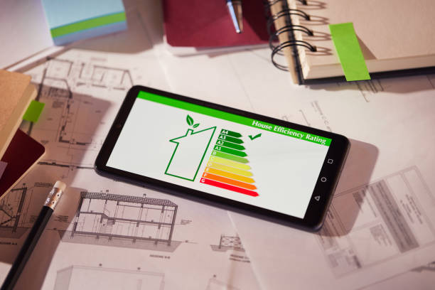 Energy efficiency screen on cellphone leaning on drawings and blueprints of a new house. Architect's desk with plans on it and a cell phone with an energy efficiency graph. Renewable energy, renovation, energy class and eco house concept. aplicación móvil de eficiencia energética en pantalla. primer plano del detalle de la calificación de eficiencia de la casa en el teléfono móvil. aplicación de lectura de eficiencia del hogar en el teléfono móvil mantenida en el escritorio.