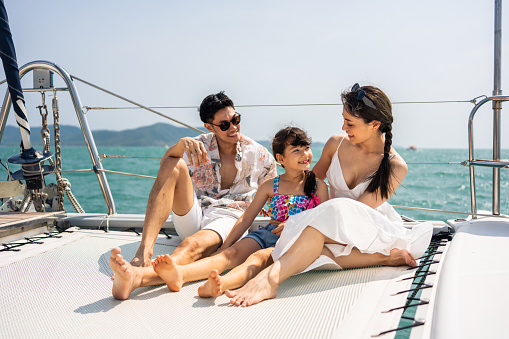 Caucasian happy family sitting on deck of yacht while yachting outdoor. Young beautiful couple hanging out and spend time with daughter while catamaran boat sailing during holiday summer trip together