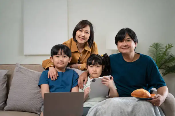 Photo of Happy parents with two kids using smartphone at home together, sitting on couch, smiling mother and father with little son and daughter looking at phone screen, having fun