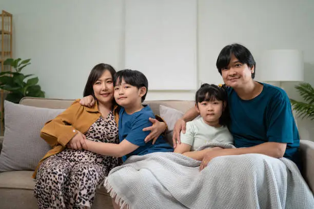 Photo of Happy parents with two kids using smartphone at home together, sitting on couch, smiling mother and father with little son and daughter looking at phone screen, having fun