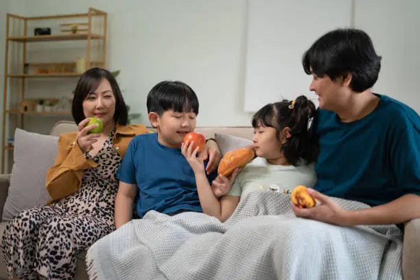 Photo of Happy parents with two kids using smartphone at home together, sitting on couch, smiling mother and father with little son and daughter looking at phone screen, having fun