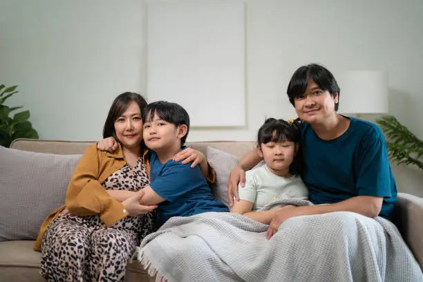 Photo of Happy parents with two kids using smartphone at home together, sitting on couch, smiling mother and father with little son and daughter looking at phone screen, having fun