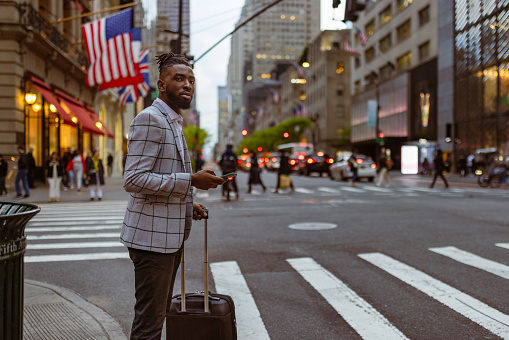 Affluent black businessman on a trip to NYC. He is walking 5th Avenue elegantly dressed with a suitcase, using mobile phone app, calling a taxi