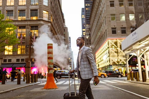 Affluent black businessman on a trip to NYC. He is walking 5th Avenue elegantly dressed with a suitcase, street crossing