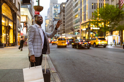 Affluent black businessman on a trip to NYC. He is walking 5th Avenue elegantly dressed with a suitcase and shopping bags in a hand calling a cab