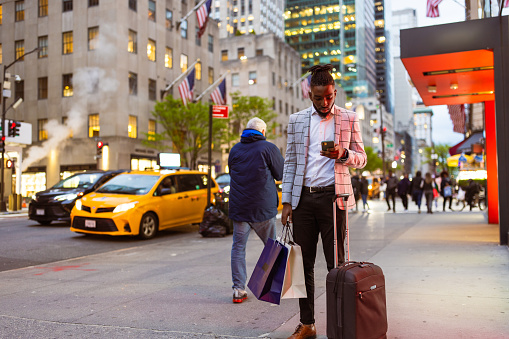 Affluent black businessman on a trip to NYC. He is walking 5th Avenue elegantly dressed with a suitcase, using mobile phone