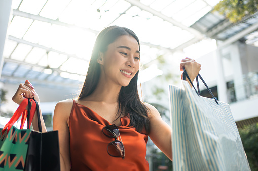 Asian beautiful woman shopping goods outdoor alone in department store. Attractive young girl holding shopping bags while walking with happiness enjoying purchasing goods in shopping mall center.