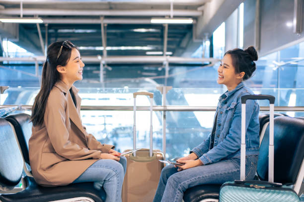 istock Asian young women talking together while waiting for airplane boarding. Attractive beautiful two girl friends feeling happy and relax, having fun communicating and discussing while sitting in airport. 1587036749