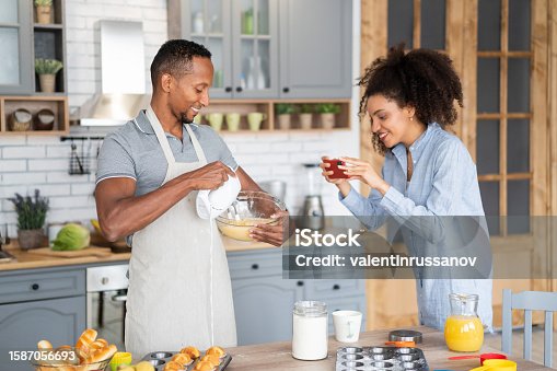 istock Couple cooking together, having fun time in the kitchen. Multiracial man mixing eggs in a bowl, while his girlfriend taking picture of him with her mobile phone. 1587056693