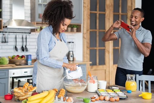 Couple cooking together, having fun time in the kitchen. Multiracial young woman mixing eggs in a bowl, while her boyfriend taking picture of her with his mobile phone, shooting home video or vlogging