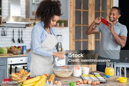 istock Couple cooking together, having fun time in the kitchen. Multiracial young woman mixing eggs in a bowl, while her boyfriend taking picture of her with his mobile phone, shooting home video or vlogging 1587056704