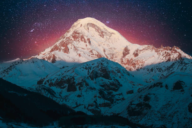 istock Georgia. Winter Bold Bright Night Starry Violet Gradient Sky With Glowing Stars Over Peak Of Mount Kazbek Covered With Snow. Amazing Night Georgian Winter Landscape. Travel To Georgia 1587062204
