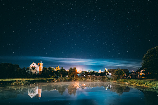 Night Clouds Over River Lake Near Houses In Village. Night Starry Sky Above Lake River With Bright Stars And Meteoric Track Trail. Glowing Stars Above Summer Nature.