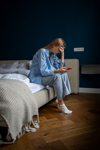 Sad unhappy young woman holding smartphone reading unpleasant sms message while sitting alone on bed, depressed female looking at phone screen receiving breakup text from boyfriend, getting bad news