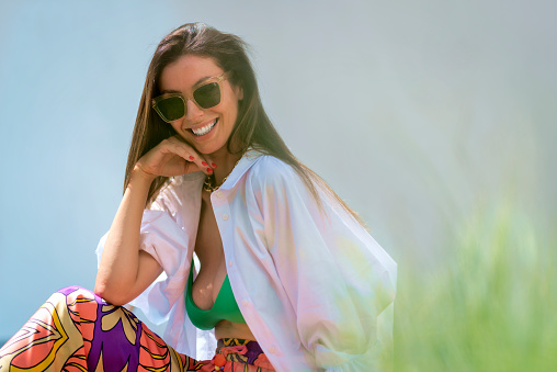 Portrait of beautiful woman with long brown hair relaxing outdoor and enjoying summer sunshine. Smiling female wearing sunglases and beachwear.
