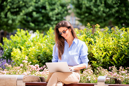 Brunette haired woman sitting on a bench in the city and using a laptop for work. Happy mid aged female wearing sunglasses as casual clothes.