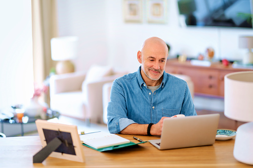 Confident senior man using notebook at home. Mid aged male working online, having video call and sitting at desk. Home office.