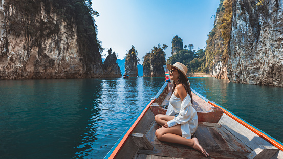 Travel summer vacation concept, Happy solo traveler asian woman with hat relax and sightseeing on Thai longtail boat in Ratchaprapha Dam at Khao Sok National Park, Surat Thani Province, Thailand