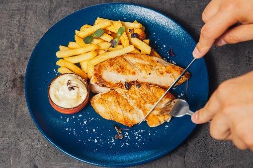 A closeup of a person cutting a chicken steak with a knife, served with fries and sauce