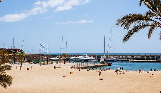 A sandy beach with palm trees and marina. The image shows a wide view of the beach, with a few people relaxing on the sand. On the left side, there are some palm trees that provide shade and contrast with the blue sky. In the background, there is a marina with several boats and yachts docked, creating a sense of luxury and leisure. The water is a light blue color and is calm, reflecting the sky and the boats. The image has a realistic and natural look, with bright colors and clear details.