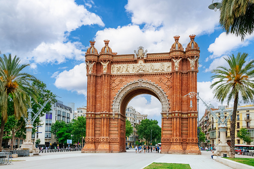 The Arc de Triomf, a monumental arch in Barcelona, Spain.The arch stands in a large plaza with palm trees and other greenery, creating a contrast between the historic and the natural. There are people walking around the plaza, enjoying the sunny day and the view of the arch. In the background, there are some buildings that show the urban landscape of Barcelona. The sky is blue with some white clouds, adding to the brightness of the scene.\nThis image can be used for travel, tourism, culture, history, architecture, or art related projects.