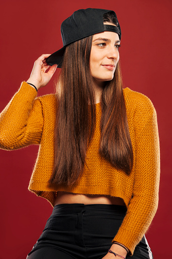 Attractive young hipster with long brunette hair in snapback and yellow sweater sitting in stool on red background at studio.