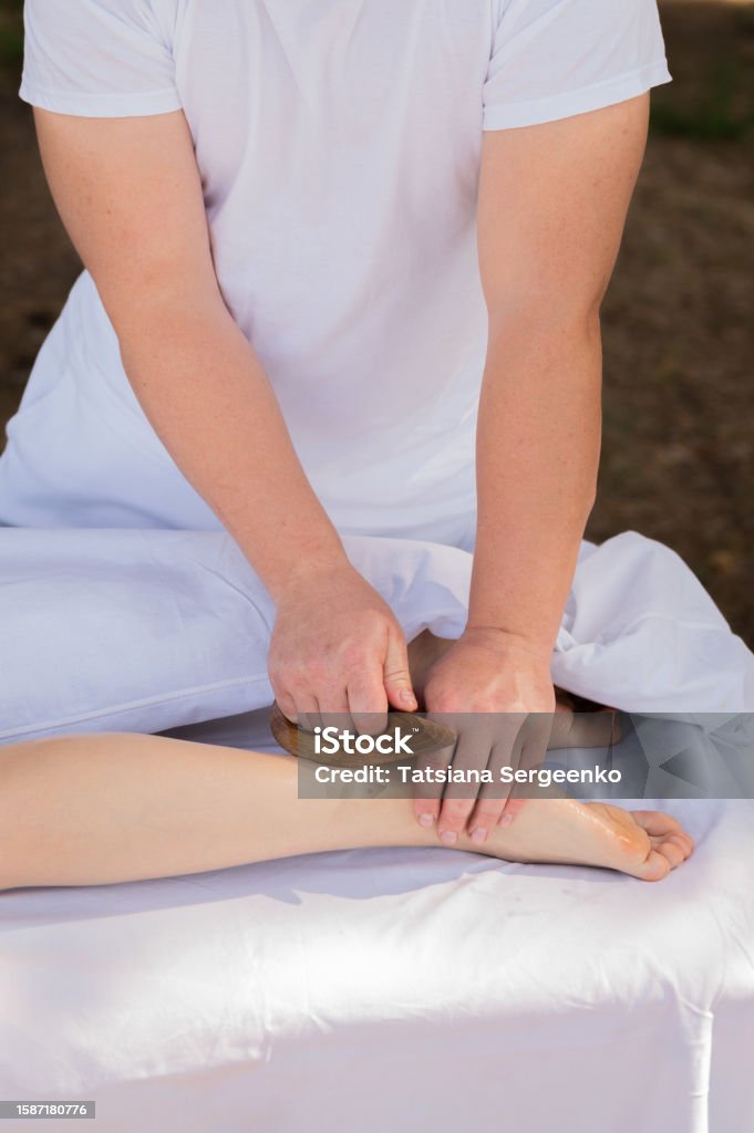 Close-up of a masseur doing foot massage with wooden tool. Lymphatic drainage massage of the back of the female leg Close-up of a masseur doing foot massage with wooden tool. Lymphatic drainage massage of the back of the female leg. Adult Stock Photo