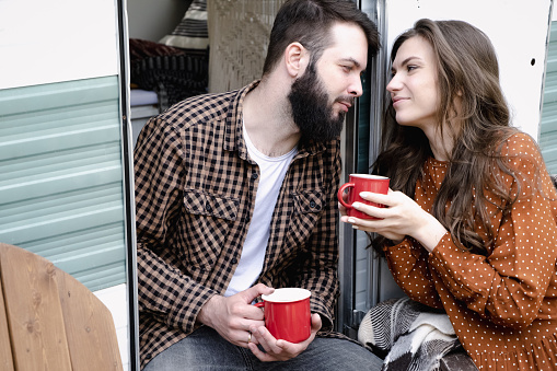 Young caucasian couple is traveling in travel van. Romantic atmosphere of relaxation. Road trip around country for the weekend. Man and woman sitting in camper and drinking tea. Millennial generation