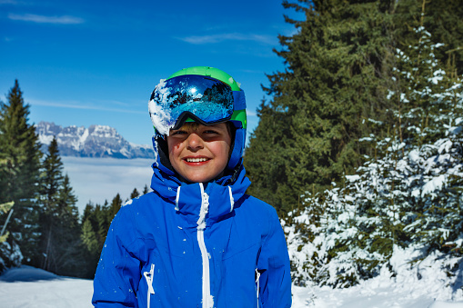 Close portrait of a laughing young boy in ski helmet and mask over snow covered mountains with fir forest