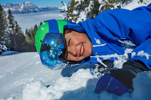 Close-up portrait of a laughing young boy in ski helmet and mask laying in snow for fun