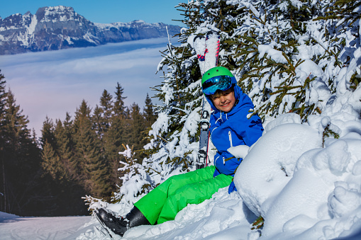 Happy young boy in ski helmet with googles and alpine skies smile sitting in snow mountains over snowy firs