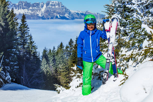 Happy young boy in ski helmet with googles and alpine skies smile stand full height in snow over snowy firs, Alps mountains