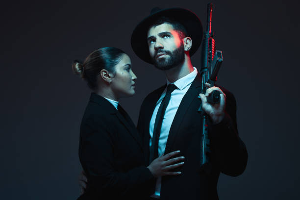 couple, fashion or weapon on dark studio background in secret spy, isolated mafia safety or crime lord security. gangster love, woman or model with gun in stylish, trendy or fashion clothes aesthetic - gun men spy suit imagens e fotografias de stock