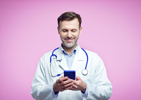 Pleased mature man wearing white lab coat and stethoscope using mobile phone. Portrait of confident male doctor against pink background.