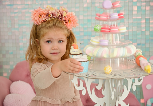 Cute little girl with a flower wreath on her head takes a cake from the festive table